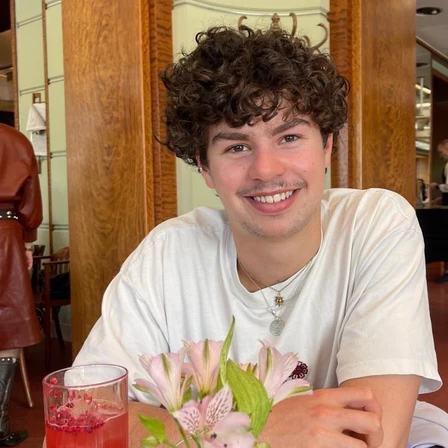 William, smiling, in a white t-shirt at a cafe table in Prague, with a cold drink and a vase of flowers.